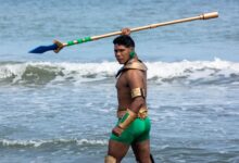 A muscular man in costume standing by the sea, holding a spear with waves in the background.