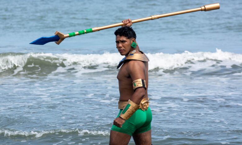 A muscular man in costume standing by the sea, holding a spear with waves in the background.