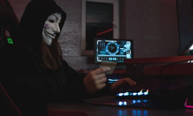 Masked hacker with credit card at computer, symbolizing cybercrime and anonymity.