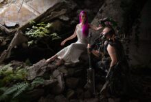 A woman in white and a sci-fi armored figure in a post-apocalyptic setting with ruins.