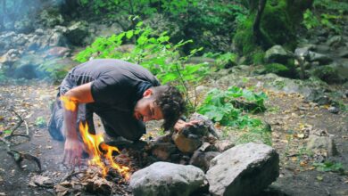 Man attempts to start a campfire surrounded by rocks in a lush green forest.