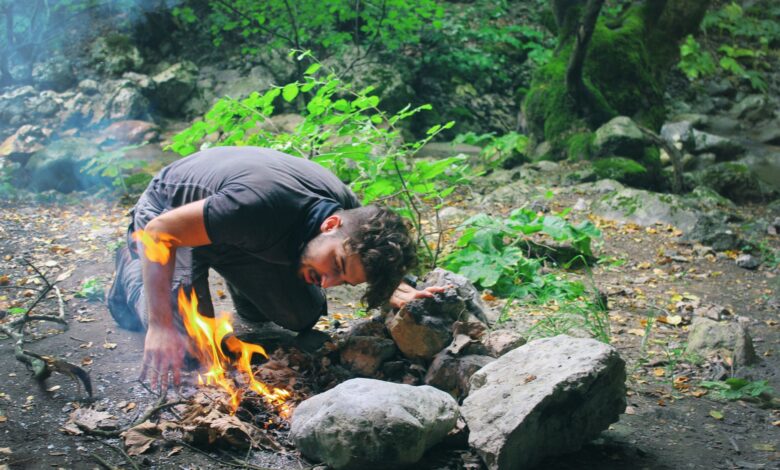 Man attempts to start a campfire surrounded by rocks in a lush green forest.