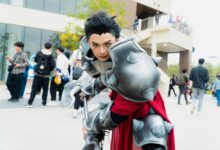 Man in intricate fantasy armor posing at a vibrant outdoor cosplay event.