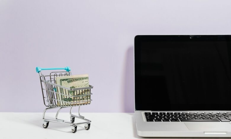 Mini shopping cart with cash beside a laptop, symbolizing online shopping and e-commerce.