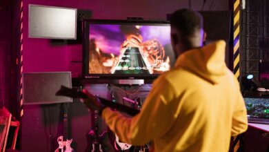 A man in a yellow jacket playing Guitar Hero on a monitor indoors, enjoying e-sports.