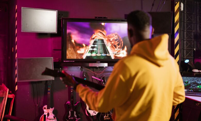 A man in a yellow jacket playing Guitar Hero on a monitor indoors, enjoying e-sports.