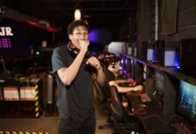 A young gamer excitedly posing in a dimly lit esports arena. Others playing in the background.