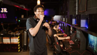 A young gamer excitedly posing in a dimly lit esports arena. Others playing in the background.