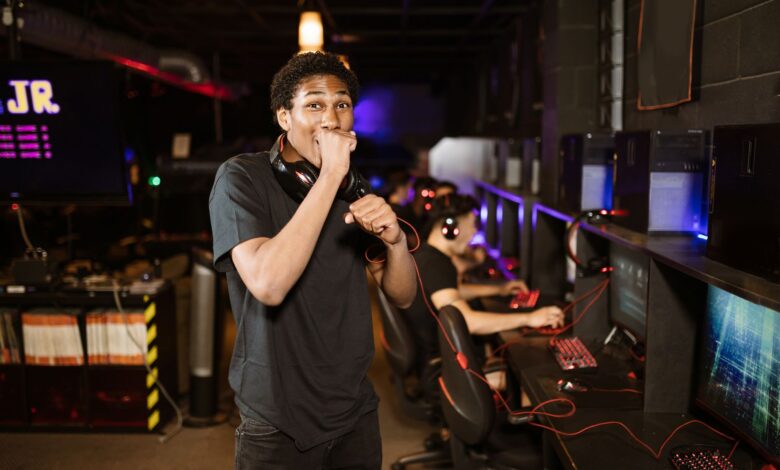 A young gamer excitedly posing in a dimly lit esports arena. Others playing in the background.