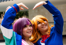 Smiling cosplayers in colorful costumes pose with a heart gesture, expressing friendship and joy.
