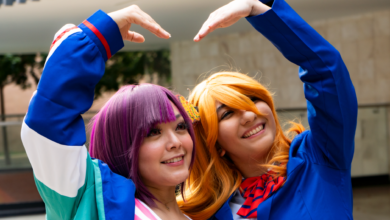 Smiling cosplayers in colorful costumes pose with a heart gesture, expressing friendship and joy.
