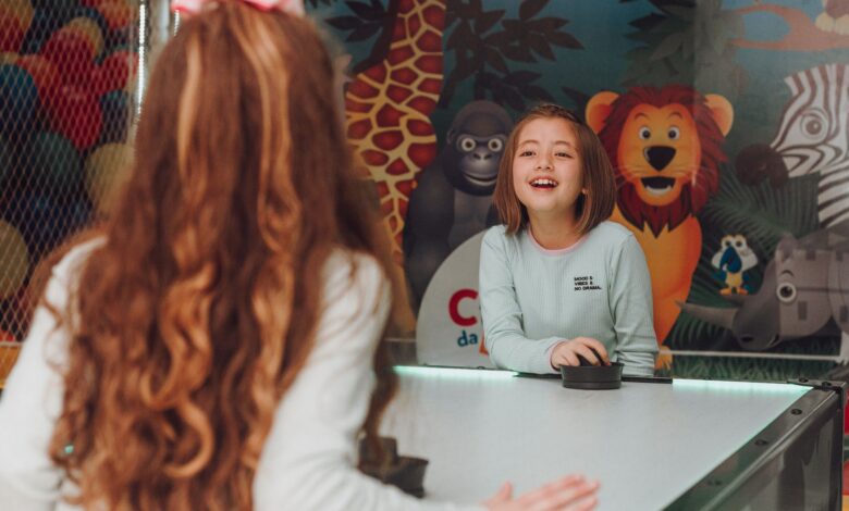 Two happy young girls playing air hockey indoors with colorful animal mural background.
