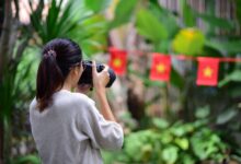 A woman takes photos in a lush outdoor setting with flags in the background.