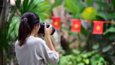 A woman takes photos in a lush outdoor setting with flags in the background.