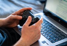 Person holds a gaming controller while interacting with a laptop, emphasizing modern gaming technology.