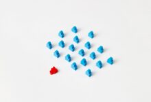 A distinctive red game piece stands out among blue meeples on a minimalist white background, symbolizing individuality.