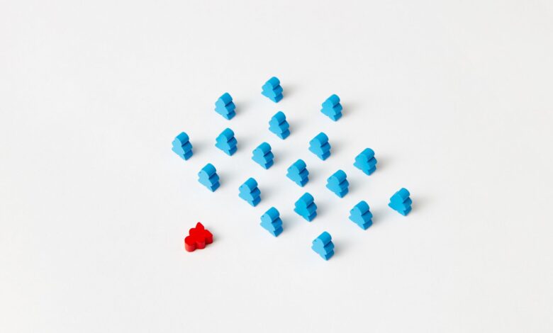 A distinctive red game piece stands out among blue meeples on a minimalist white background, symbolizing individuality.