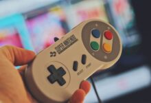 Hand holding a classic Super Nintendo game controller indoors with a blurred screen in the background.
