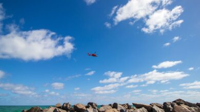 Vivid image of a red helicopter flying over rocky coastline in Miami under a bright blue sky.