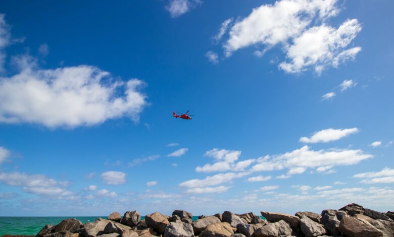 Vivid image of a red helicopter flying over rocky coastline in Miami under a bright blue sky.