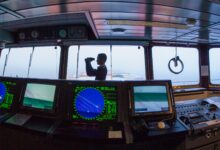 Silhouette of a navigator using binoculars on a cargo ship's bridge, surrounded by navigation equipment.