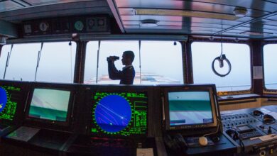 Silhouette of a navigator using binoculars on a cargo ship's bridge, surrounded by navigation equipment.