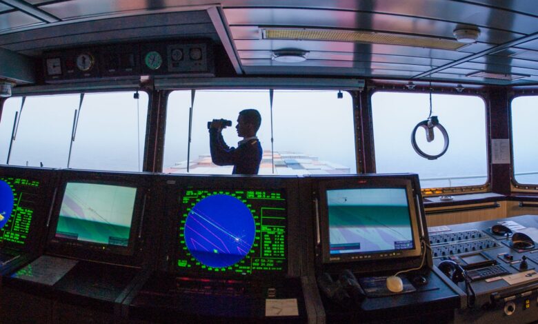 Silhouette of a navigator using binoculars on a cargo ship's bridge, surrounded by navigation equipment.