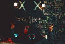 A masked individual stands before a chalkboard filled with neon drawings and cryptic messages, creating a mysterious ambiance.
