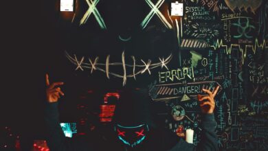 A masked individual stands before a chalkboard filled with neon drawings and cryptic messages, creating a mysterious ambiance.
