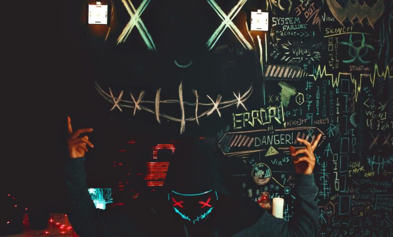 A masked individual stands before a chalkboard filled with neon drawings and cryptic messages, creating a mysterious ambiance.