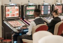 Two men engaged in gaming at a casino, seated at slot machines indoors.