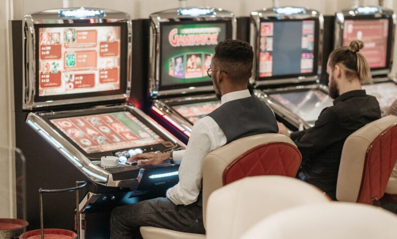 Two men engaged in gaming at a casino, seated at slot machines indoors.