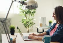 Woman smiling while working on a laptop in a modern office with plants.