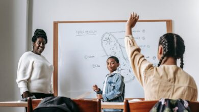 Ethnic girl raising hand while African American female teacher standing near whiteboard with teen boy and explaining task