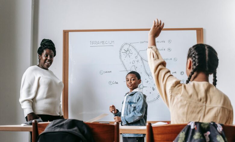 Ethnic girl raising hand while African American female teacher standing near whiteboard with teen boy and explaining task