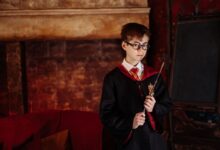 Boy dressed as a wizard with eyeglasses and robe holding a wand in front of a fireplace.