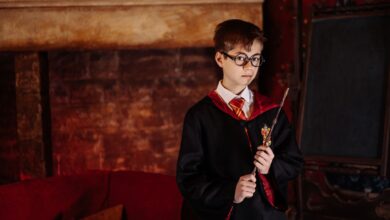 Boy dressed as a wizard with eyeglasses and robe holding a wand in front of a fireplace.