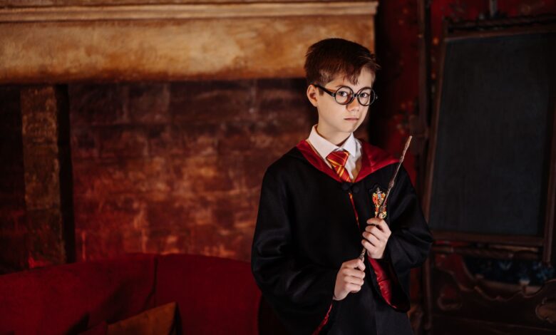 Boy dressed as a wizard with eyeglasses and robe holding a wand in front of a fireplace.