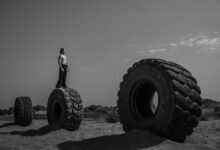 Black and white image of man standing on huge tires in a barren desert setting, suggesting resilience.
