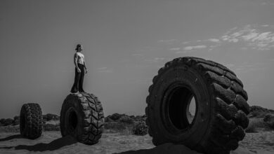 Black and white image of man standing on huge tires in a barren desert setting, suggesting resilience.