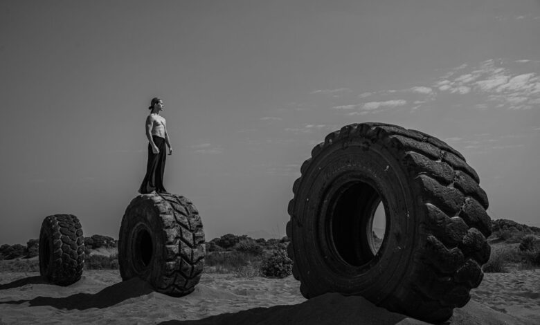 Black and white image of man standing on huge tires in a barren desert setting, suggesting resilience.
