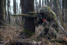 Sniper in camouflage gear taking aim in a dense forest environment.