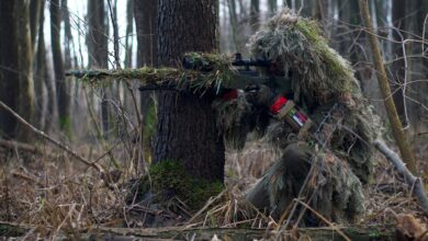 Sniper in camouflage gear taking aim in a dense forest environment.