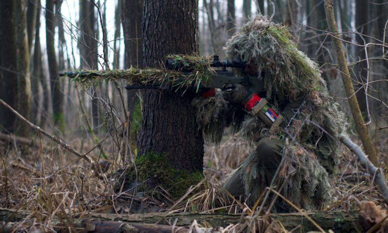 Sniper in camouflage gear taking aim in a dense forest environment.