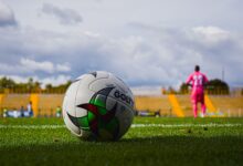 Close-up of a soccer ball on green grass field with a blurred player in pink jersey in the background during a match.