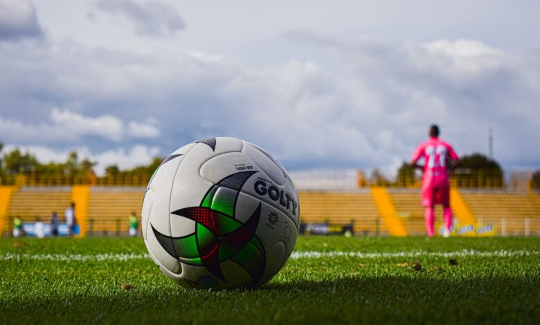 Close-up of a soccer ball on green grass field with a blurred player in pink jersey in the background during a match.