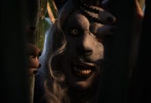 A frightening costumed figure peers through shadows with glowing eyes and sharp teeth, evoking terror in a dark, mysterious setting.