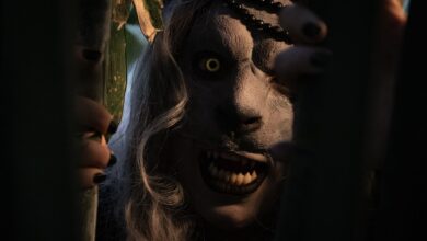 A frightening costumed figure peers through shadows with glowing eyes and sharp teeth, evoking terror in a dark, mysterious setting.