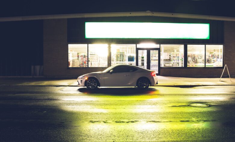 White coupe car on roadside at glowing shop with signboard late at night time