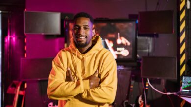 Smiling gamer in a yellow hoodie stands confidently in an e-sports studio with vibrant lighting.